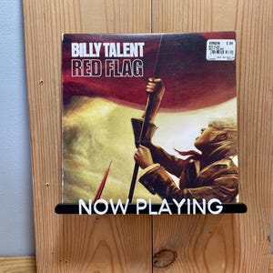 May include: A vinyl record by Billy Talent titled "Red Flag" is displayed on a wooden wall shelf with a black "NOW PLAYING" sign.
