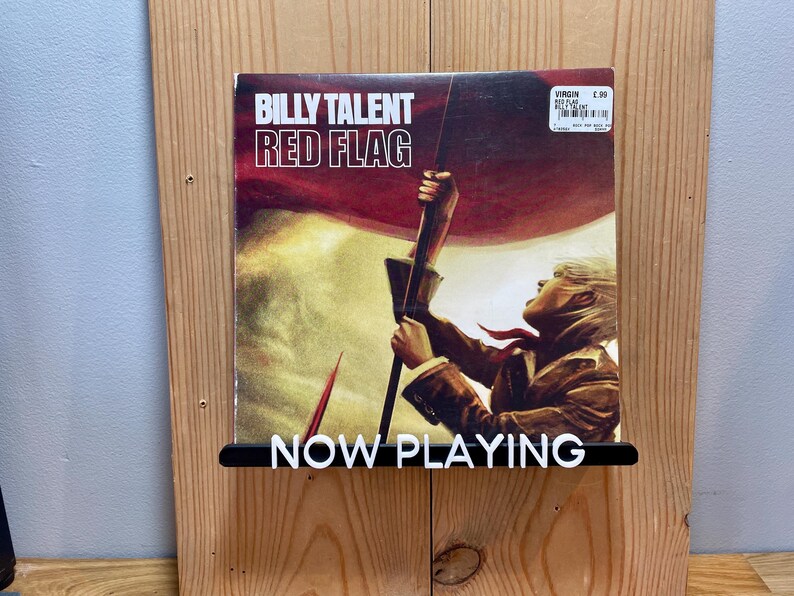 May include: A vinyl record by Billy Talent titled "Red Flag" is displayed on a wooden wall shelf with a black "NOW PLAYING" sign.