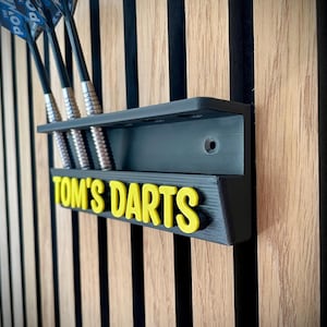 以下が含まれることがあります： 「Tom's Darts」の文字が黄色で書かれた、黒い壁掛けダーツホルダー。ホルダーにはダーツ用のスロットがあり、4本のダーツが見えます。ホルダーは、縦縞模様の木製の壁に取り付けられています。
