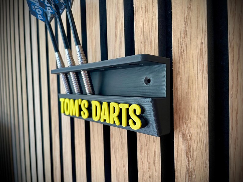 以下が含まれることがあります： 「Tom's Darts」の文字が黄色で書かれた、黒い壁掛けダーツホルダー。ホルダーにはダーツ用のスロットがあり、4本のダーツが見えます。ホルダーは、縦縞模様の木製の壁に取り付けられています。