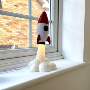 Op de afbeelding: Een decoratieve raketlamp in wit en rood, met een zwart patrijspoort en vinnen. De lamp straalt warm licht uit vanaf de basis en staat op een witte vensterbank. Links staat een kleine vetplant.