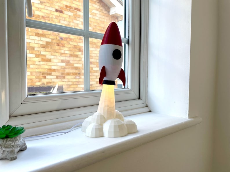 Op de afbeelding: Een decoratieve raketlamp in wit en rood, met een zwart patrijspoort en vinnen. De lamp straalt warm licht uit vanaf de basis en staat op een witte vensterbank. Links staat een kleine vetplant.