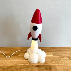 Op de afbeelding: Een decoratieve raketvormige lamp met een rode kegel, witte behuizing en zwarte accenten. De lamp heeft rode vinnen en een wolkvormige basis, met licht dat van onderaf komt. Het is verbonden met een netsnoer met een aan/uit-schakelaar.