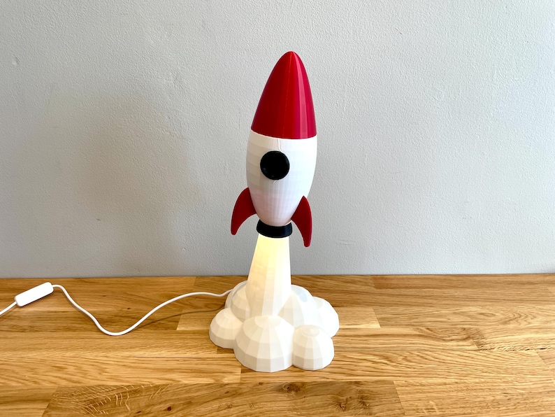 Op de afbeelding: Een decoratieve raketvormige lamp met een rode kegel, witte behuizing en zwarte accenten. De lamp heeft rode vinnen en een wolkvormige basis, met licht dat van onderaf komt. Het is verbonden met een netsnoer met een aan/uit-schakelaar.
