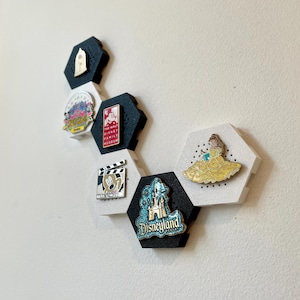 May include: A collection of Disney pins displayed on a wall. The pins are attached to hexagonal black and white holders. The pins feature various Disney characters and logos, including a Disneyland pin and a Belle pin.