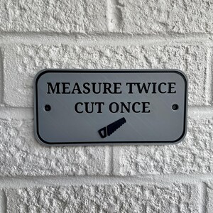 May include: A rectangular, silver-colored sign with rounded corners is mounted on a white brick wall. The sign features the text "MEASURE TWICE CUT ONCE" in black, along with a saw icon.