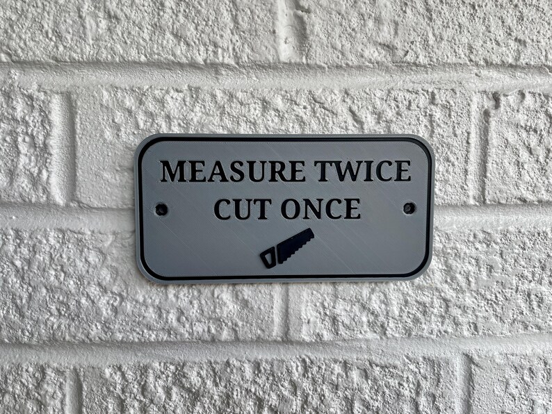 May include: A rectangular, silver-colored sign with rounded corners is mounted on a white brick wall. The sign features the text "MEASURE TWICE CUT ONCE" in black, along with a saw icon.