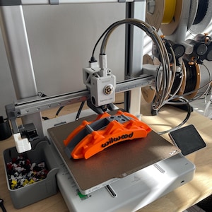 May include: A 3D printer with an orange Wilwood part on the print bed. The printer is gray and white, with multiple spools of filament in the background. The text "PRINT EASTWOOD" is visible above the printer.