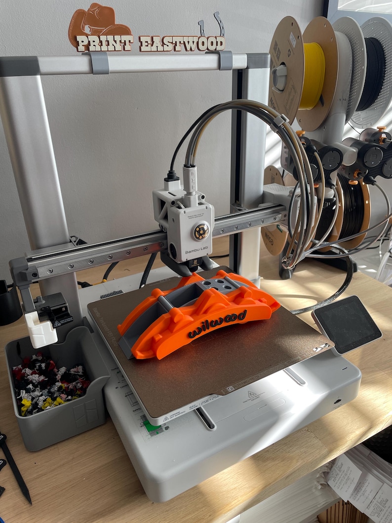 May include: A 3D printer with an orange Wilwood part on the print bed. The printer is gray and white, with multiple spools of filament in the background. The text "PRINT EASTWOOD" is visible above the printer.