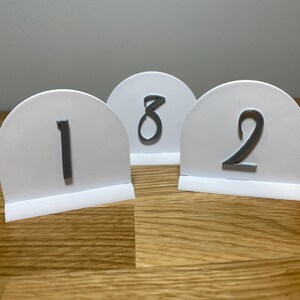 May include: Three white table number holders with gray numbers 1, 2, and 8. The numbers are printed on a rounded, white surface with a base.