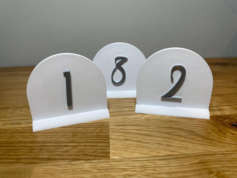 May include: Three white table number holders with gray numbers 1, 2, and 8. The numbers are printed on a rounded, white surface with a base.