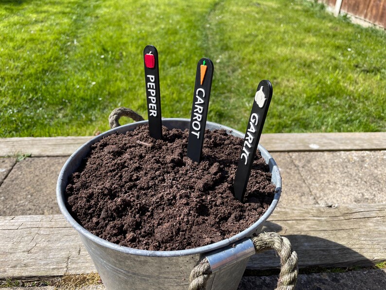May include: Three black plant markers with white text and colorful icons. The markers are labeled "Pepper", "Carrot", and "Garlic". The markers are in a metal bucket filled with dark brown soil.