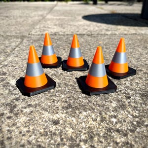 May include: Five orange and grey traffic cones with black bases are arranged on a textured concrete surface. The cones have a reflective grey band and are designed for road safety and construction.