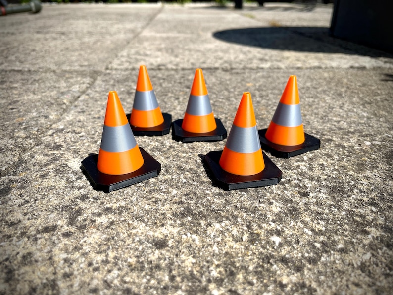 May include: Five orange and grey traffic cones with black bases are arranged on a textured concrete surface. The cones have a reflective grey band and are designed for road safety and construction.