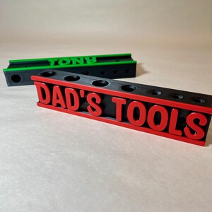 May include: Black and red tool organizer with the text "DAD'S TOOLS" in red. The organizer has several round holes for tool storage. A green and black organizer with the name "TONY" is in the background.