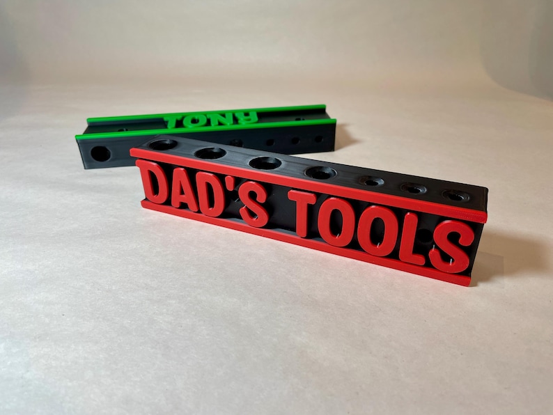 May include: Black and red tool organizer with the text "DAD'S TOOLS" in red. The organizer has several round holes for tool storage. A green and black organizer with the name "TONY" is in the background.