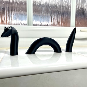 May include: A black 3D printed model of a sea serpent with googly eyes. The serpent is in three pieces, with the head and tail standing upright and the body forming a curved arch.