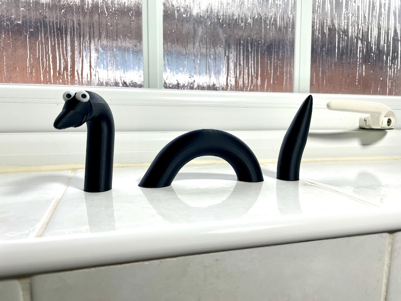 May include: A black 3D printed model of a sea serpent with googly eyes. The serpent is in three pieces, with the head and tail standing upright and the body forming a curved arch.