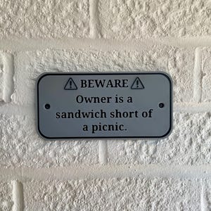 May include: A rectangular, silver-colored sign with rounded corners is mounted on a white brick wall. The sign has black text that reads "BEWARE" above "Owner is a sandwich short of a picnic."