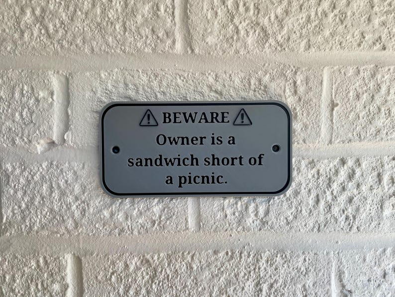 May include: A rectangular, silver-colored sign with rounded corners is mounted on a white brick wall. The sign has black text that reads "BEWARE" above "Owner is a sandwich short of a picnic."