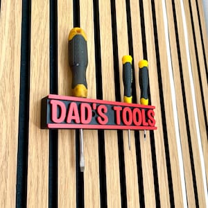 May include: A wall-mounted tool holder with the text "DAD'S TOOLS" in red letters. Three screwdrivers are inserted into the holder. The holder is black with a red border. The background is a wooden wall with vertical stripes.