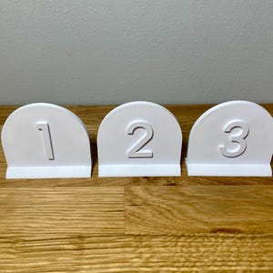 Wedding table numbers in white standing up on a wooden desk with grey background. Numbers 1 2 3 shown