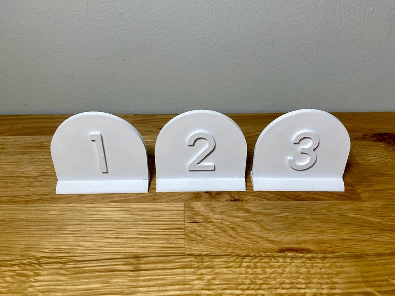 Wedding table numbers in white standing up on a wooden desk with grey background. Numbers 1 2 3 shown