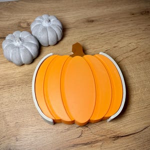 Puede incluir: Una decoraci&oacute;n en forma de calabaza naranja con un tallo marr&oacute;n y un borde blanco. Tambi&eacute;n hay dos peque&ntilde;as calabazas grises. Los art&iacute;culos est&aacute;n sobre una superficie de madera, evocando un tema de oto&ntilde;o o cosecha.