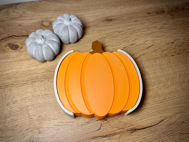 Puede incluir: Una decoraci&oacute;n en forma de calabaza naranja con un tallo marr&oacute;n y un borde blanco. Tambi&eacute;n hay dos peque&ntilde;as calabazas grises. Los art&iacute;culos est&aacute;n sobre una superficie de madera, evocando un tema de oto&ntilde;o o cosecha.