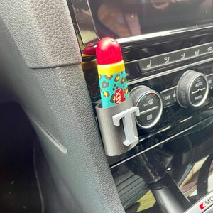 Pu&ograve; includere: Un interno di un'auto grigio con un portaoggetti grigio e bianco con la lettera "T" su di esso. Un tubo di balsamo per labbra con un design a stampa leopardata &egrave; nel portaoggetti.