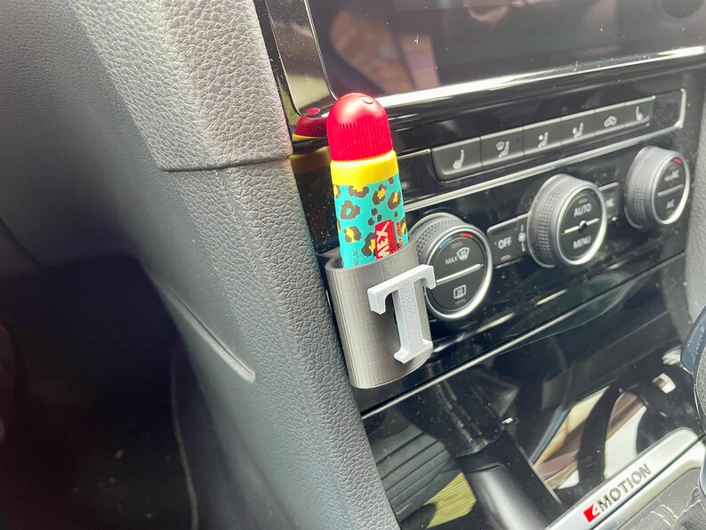 Pu&ograve; includere: Un interno di un'auto grigio con un portaoggetti grigio e bianco con la lettera "T" su di esso. Un tubo di balsamo per labbra con un design a stampa leopardata &egrave; nel portaoggetti.