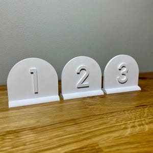 Wedding table numbers in white standing up on a wooden desk with grey background. Numbers 1 2 3 shown