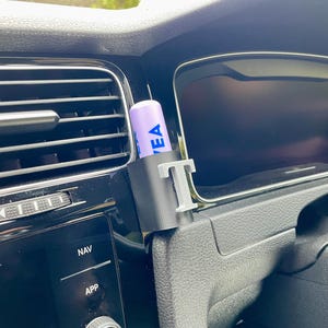 Pu&ograve; includere: Un supporto per telefono da auto stampato in 3D di colore grigio con la lettera "T" stampata su di esso. Un balsamo per labbra viola con la parola "EA" stampata su di esso &egrave; nel supporto. Il supporto &egrave; attaccato alla ventola di un'auto.