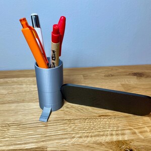 pen pot on the right with a sign that says check in here. Shown on a wooden desk with grey background. Silver pen pot with a black sign with silver text.
