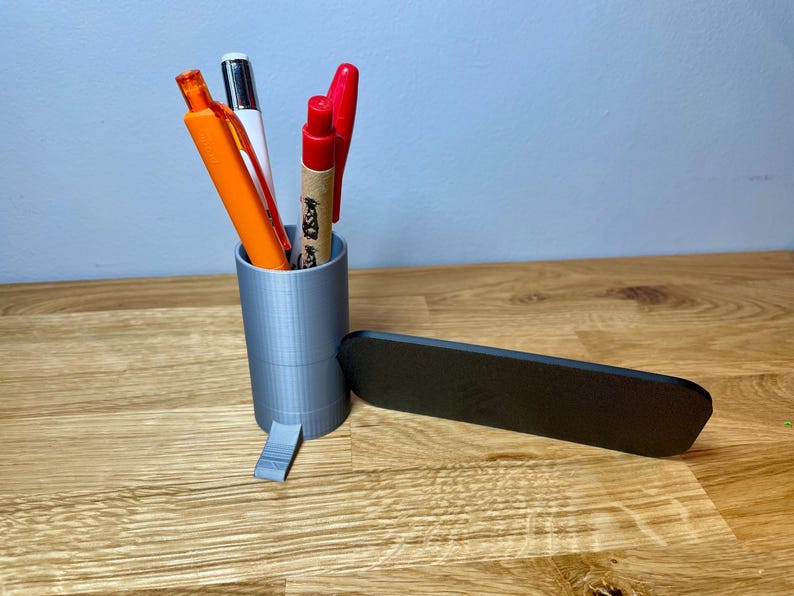 pen pot on the right with a sign that says check in here. Shown on a wooden desk with grey background. Silver pen pot with a black sign with silver text.