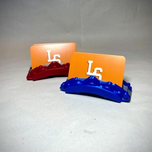 May include: Two orange card holders with white "LS" lettering, each resting on a colored base. One base is red, the other is blue. The card holders are displayed on a light gray surface.