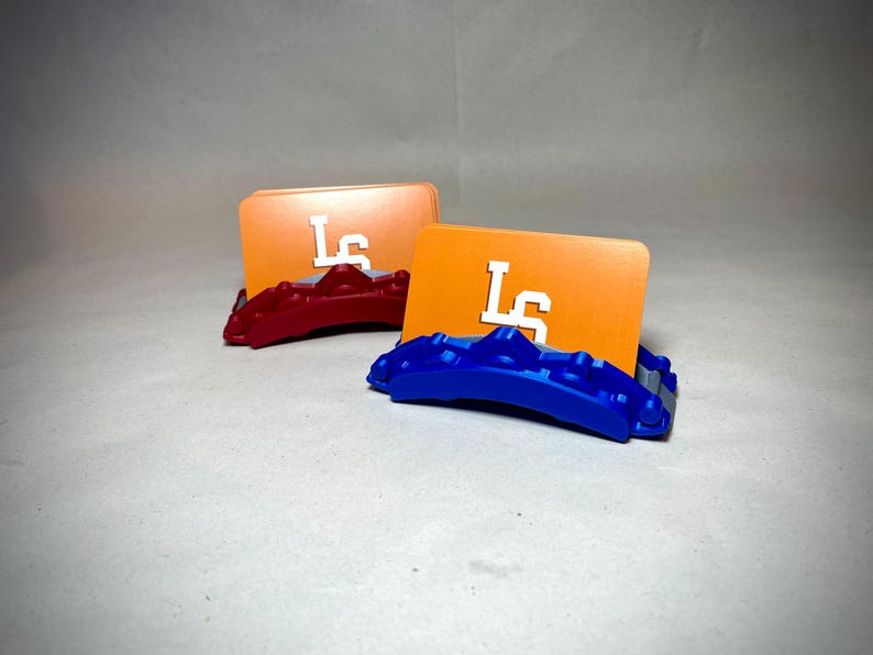 May include: Two orange card holders with white "LS" lettering, each resting on a colored base. One base is red, the other is blue. The card holders are displayed on a light gray surface.