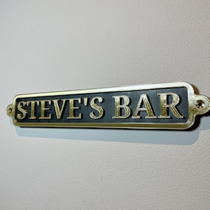 May include: A gold-colored metal sign with rounded edges and black lettering that reads "STEVE'S BAR".