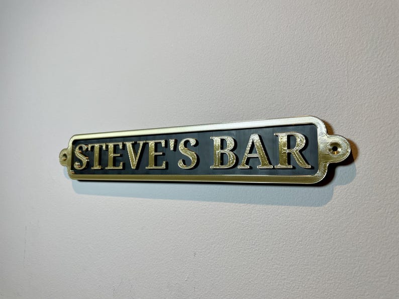 May include: A gold-colored metal sign with rounded edges and black lettering that reads "STEVE'S BAR".