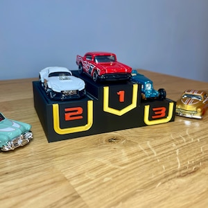 May include: A black three-tiered display stand with yellow outlines and orange numbers 1, 2, and 3. A red car sits on the top tier, a white car on the second, and a blue car on the third. A light green car and a gold car are placed on the surface.