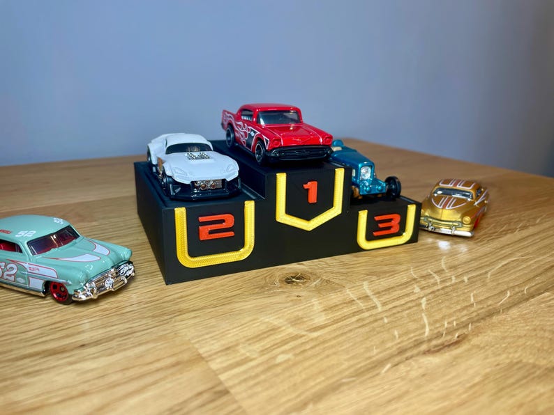 May include: A black three-tiered display stand with yellow outlines and orange numbers 1, 2, and 3. A red car sits on the top tier, a white car on the second, and a blue car on the third. A light green car and a gold car are placed on the surface.