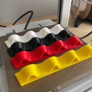 May include: Four wavy, 3D-printed holders in white, black, red, and yellow. The holders are arranged on a gray surface, likely a 3D printer bed. The design features a series of rounded peaks and valleys, suggesting a functional or decorative purpose.
