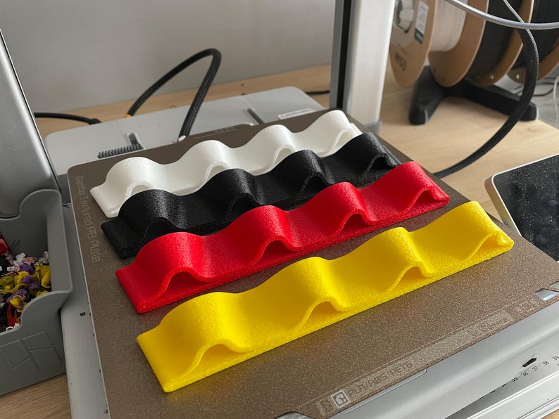 May include: Four wavy, 3D-printed holders in white, black, red, and yellow. The holders are arranged on a gray surface, likely a 3D printer bed. The design features a series of rounded peaks and valleys, suggesting a functional or decorative purpose.
