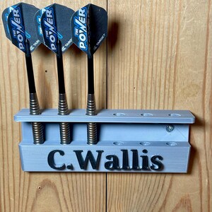 Silver / grey wall mounted dart holder with large black text along the lower edge showing a name. 3 darts in the holder with a total of 6 available slots. Shown mounted to a wooden background.