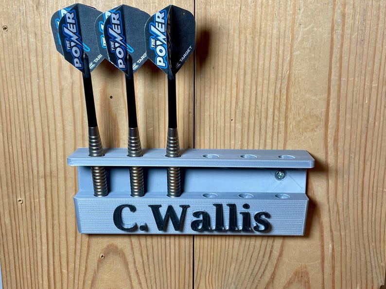 Silver / grey wall mounted dart holder with large black text along the lower edge showing a name. 3 darts in the holder with a total of 6 available slots. Shown mounted to a wooden background.