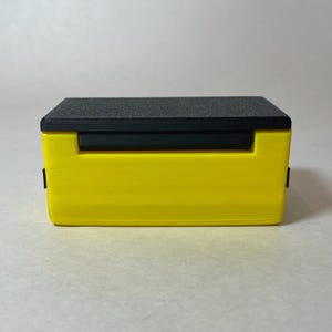Pu&ograve; includere: Una scatola rettangolare gialla con un coperchio nero e una barra orizzontale nera. La parte superiore ha una superficie strutturata grigio scuro. La scatola ha un design semplice e moderno e sembra essere di plastica.
