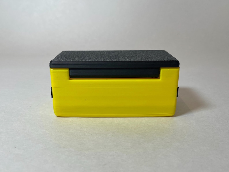 Pu&ograve; includere: Una scatola rettangolare gialla con un coperchio nero e una barra orizzontale nera. La parte superiore ha una superficie strutturata grigio scuro. La scatola ha un design semplice e moderno e sembra essere di plastica.