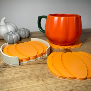 Puede incluir: Una taza naranja con forma de calabaza con un asa verde se encuentra sobre un posavasos con forma de calabaza. Un juego de posavasos de calabaza naranja est&aacute; en un soporte blanco. Tambi&eacute;n hay figuras decorativas de calabazas blancas y grises.
