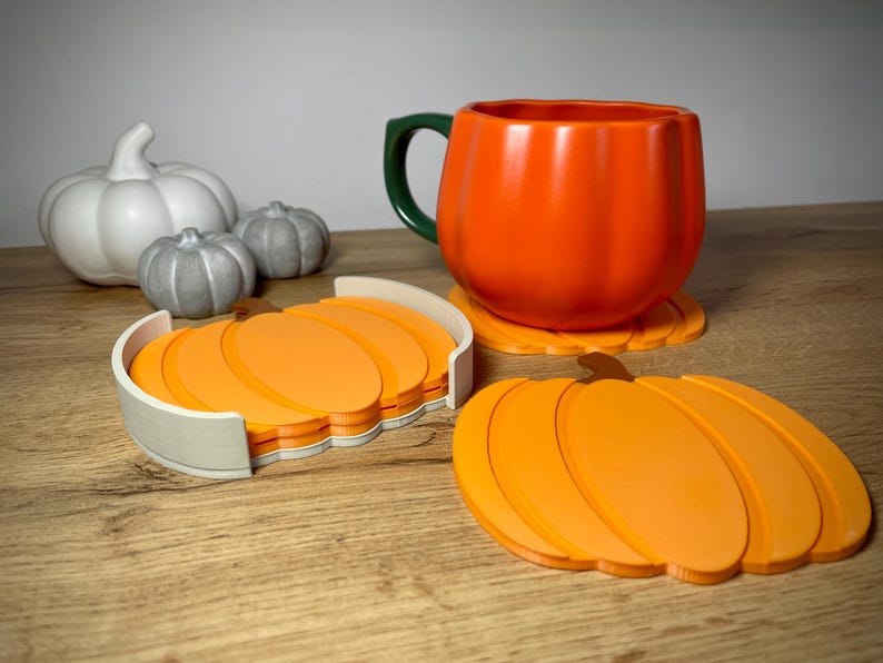 Puede incluir: Una taza naranja con forma de calabaza con un asa verde se encuentra sobre un posavasos con forma de calabaza. Un juego de posavasos de calabaza naranja est&aacute; en un soporte blanco. Tambi&eacute;n hay figuras decorativas de calabazas blancas y grises.