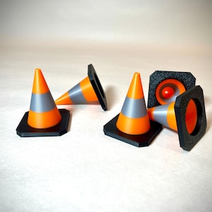 May include: A collection of small orange and grey traffic cones with black bases, set against a light backdrop. Each cone features a reflective grey band, designed to mimic road safety equipment. The cones are a bright orange colour.